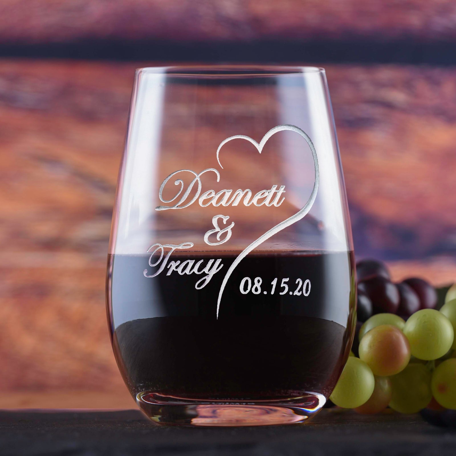 Stemless wine glass filled with dark wine, deep-etched with a heart design around the names "Deanett & Tracy" and the date "08.15.20," on a wood background with grapes.