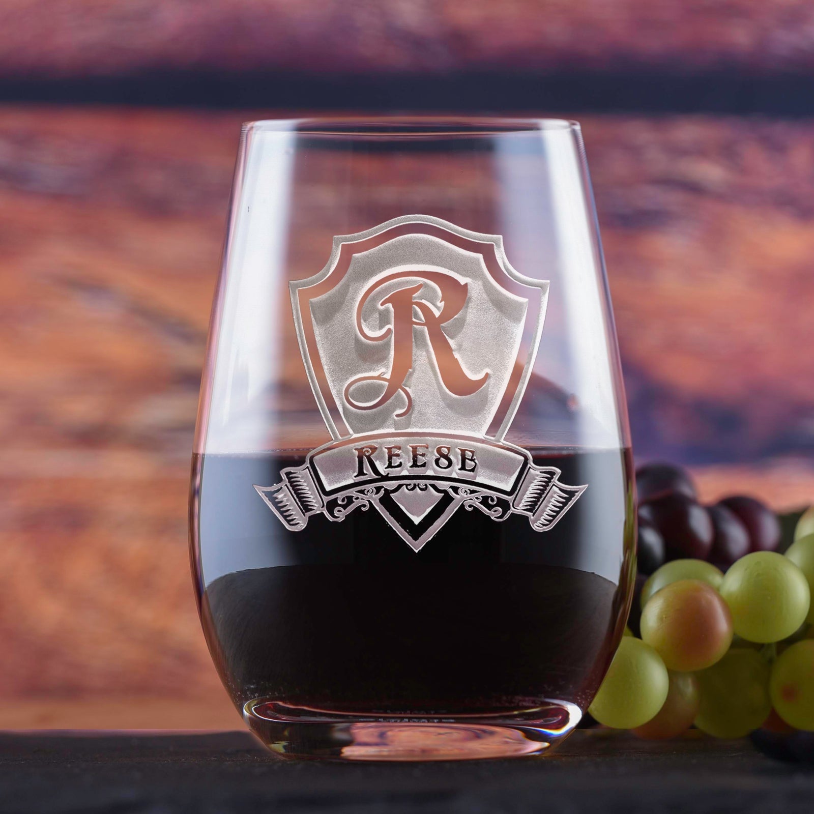 Stemless wine glass deep-etched with a shield monogram reading "R, Reese," filled with red wine, on a blurred wood-toned background.