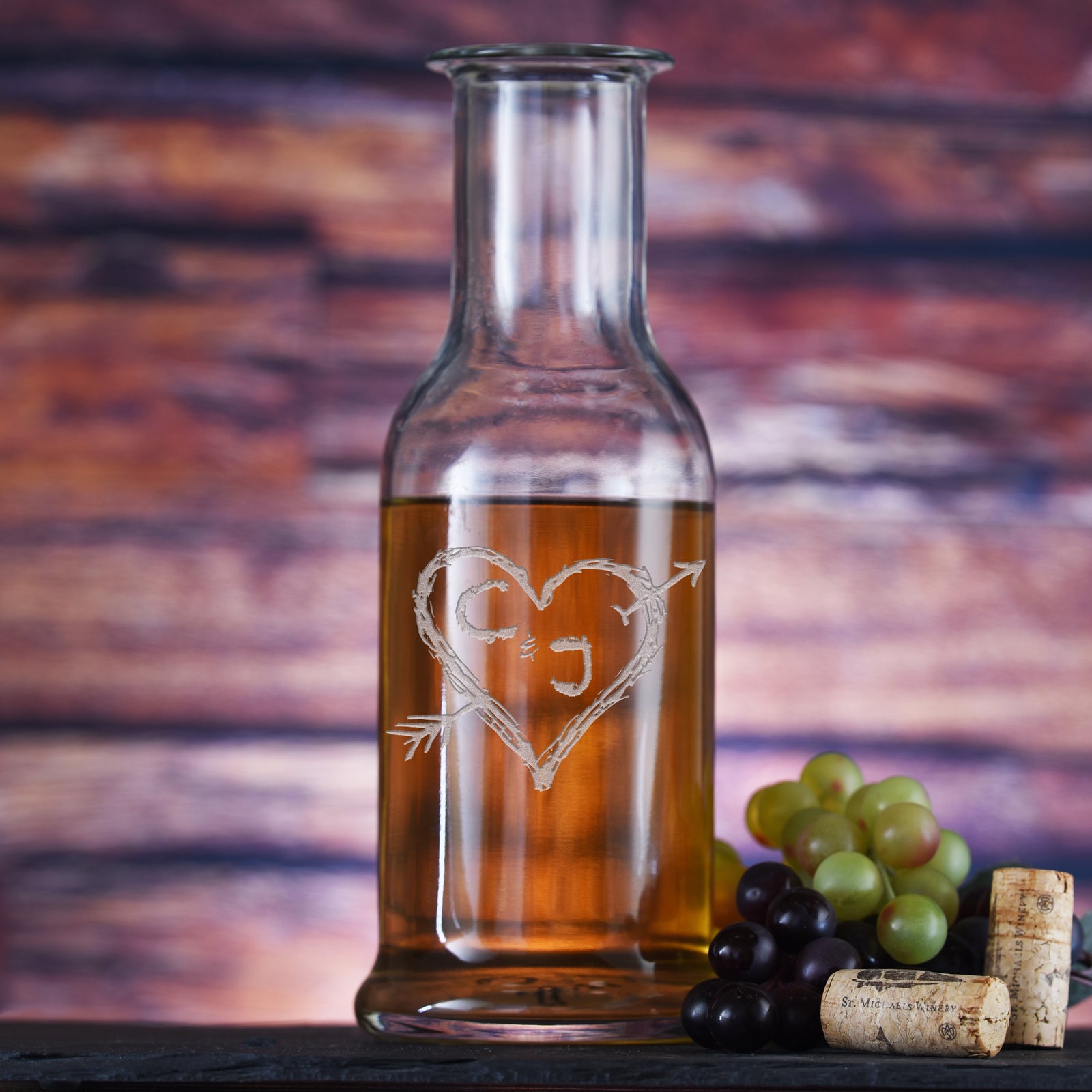 Round-shoulder wine carafe filled with white wine, deep-carved with the initials "C" and "J" inside a rustic heart pierced by an arrow, styled with grapes.