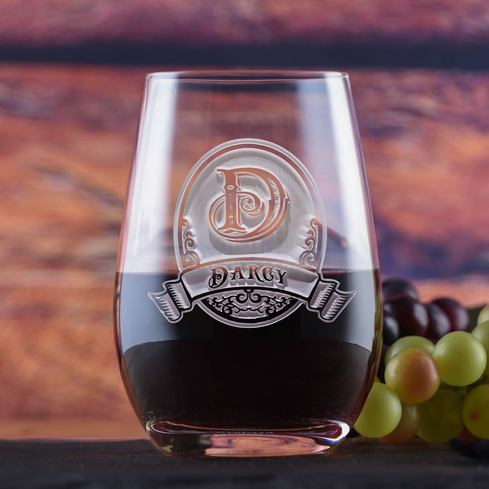 Clear stemless wine glass filled with red wine, deep-etched with an ornate oval monogram letter "D" and a banner reading "Darcy," on a blurred wood-toned background.