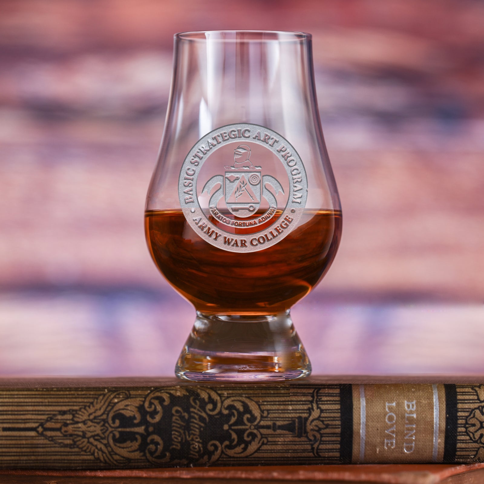 Tulip-shaped nosing glass filled with amber liquid, deep-carved with the Basic Strategic Art Program, Army War College seal (a knight's helmet above a shield with wings, reading "Paratos Fortuna Adiuvat"), resting on a book.