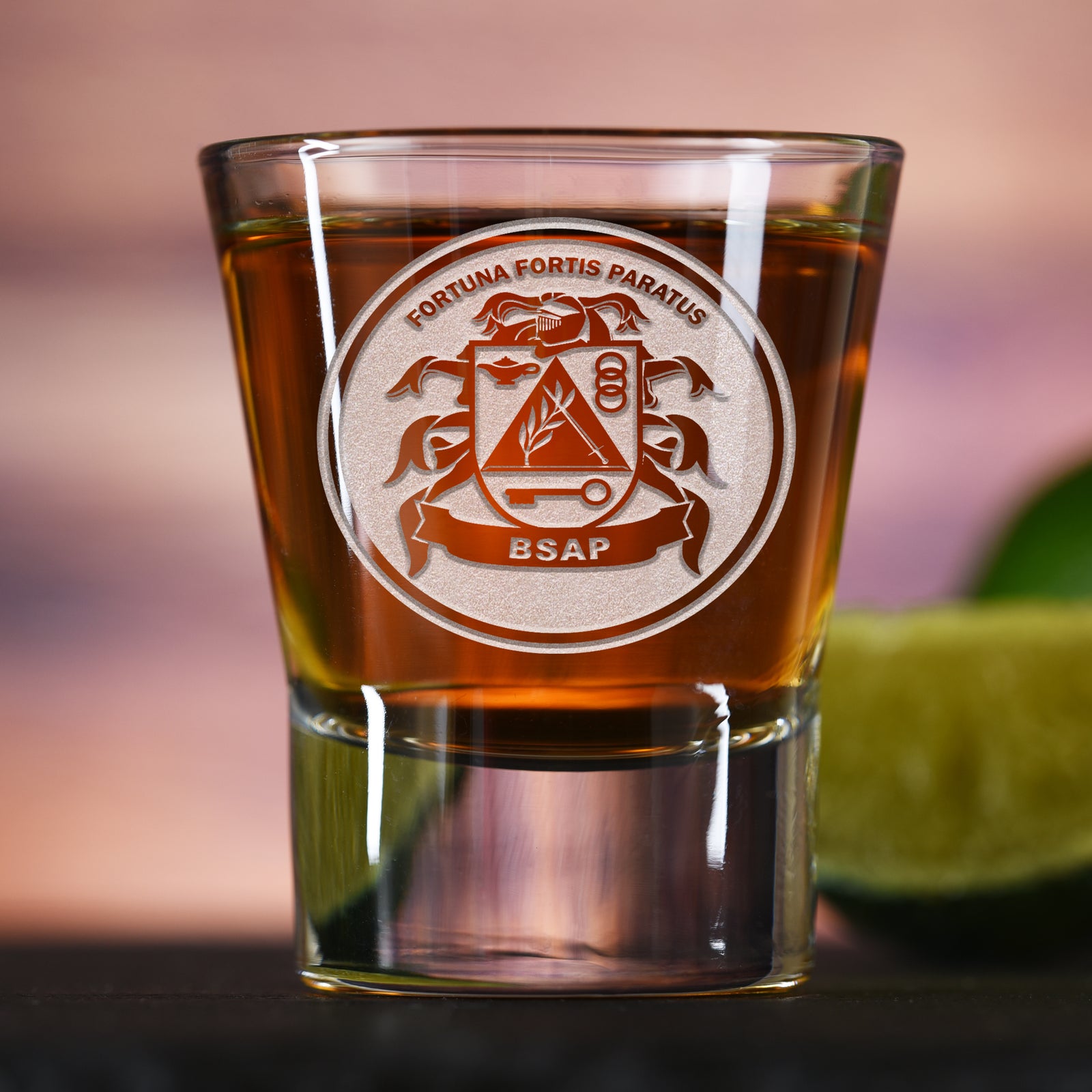 Shot glass filled with amber liquid, deep-carved with a crest reading "Fortuna Fortis Paratus, BSAP," on a bar counter with a lime.