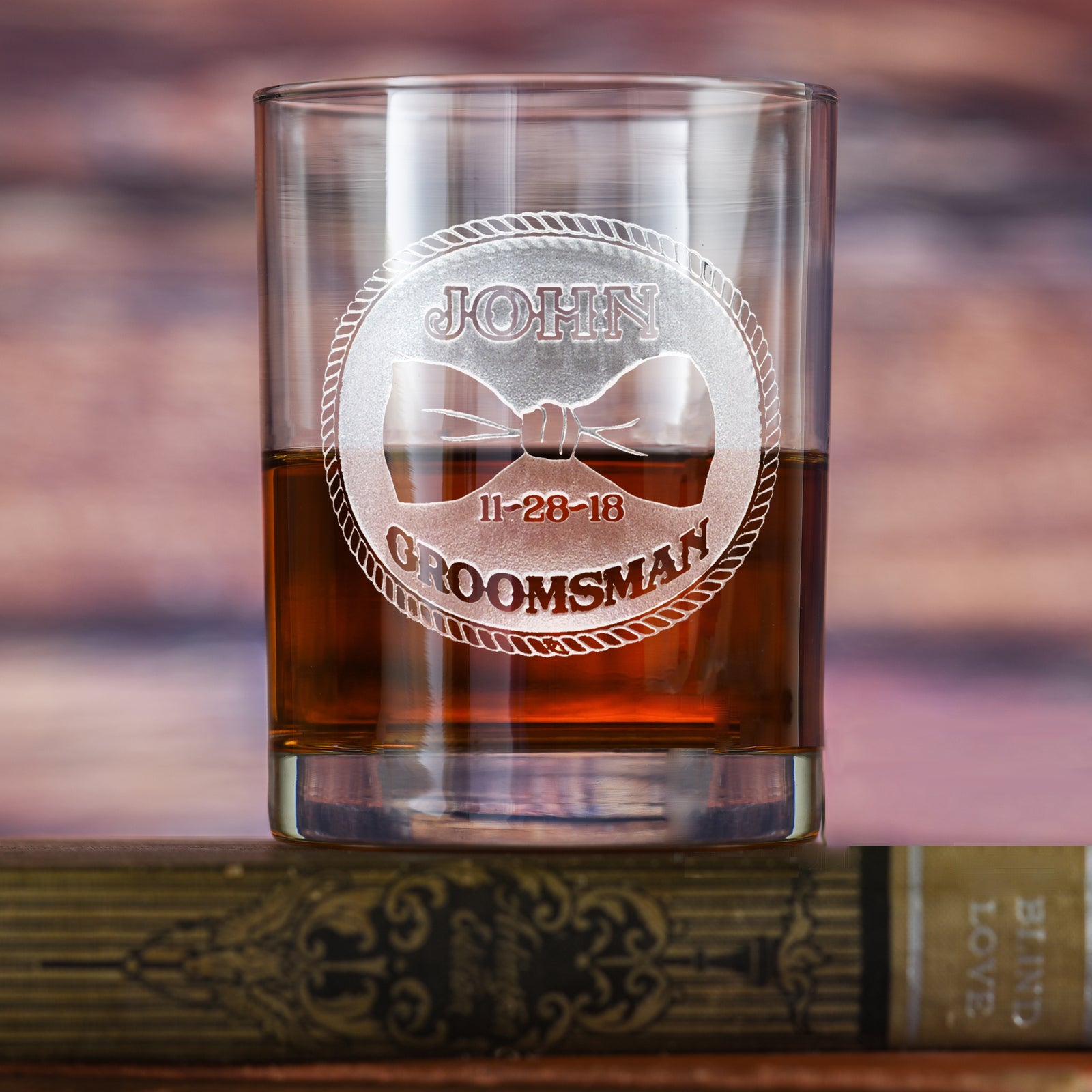 Rocks glass filled with amber liquid, deep-carved with a circular badge reading "John, Groomsman, 11-28-18" around a bowtie, resting on a book with a cigar.