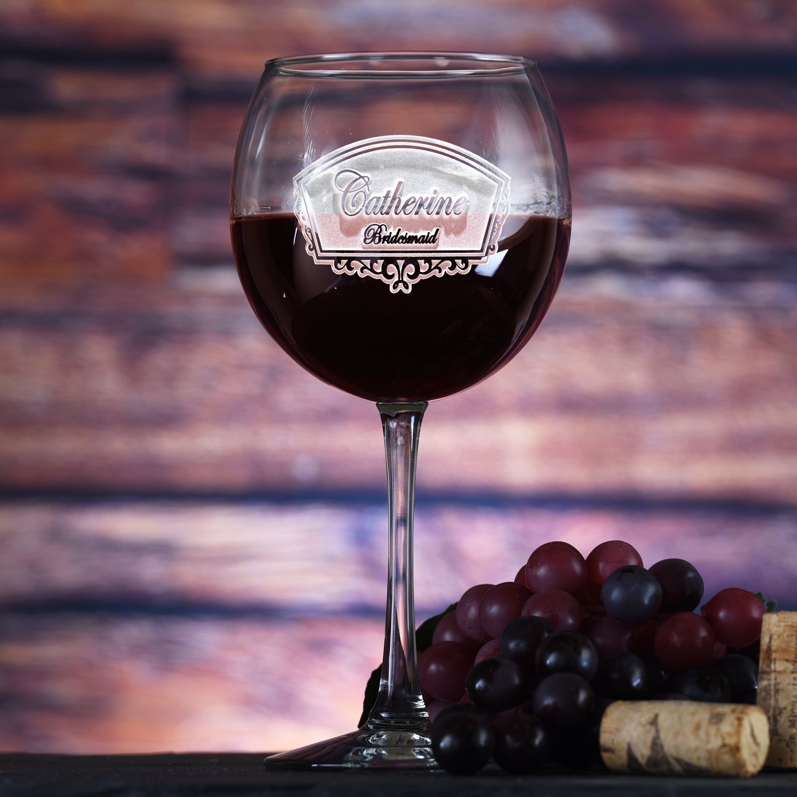 Red wine glass filled with red wine, engraved with a framed script-text plaque reading "Catherine, Bridesmaid," on a sunset-toned wood background with grapes and a wine cork.