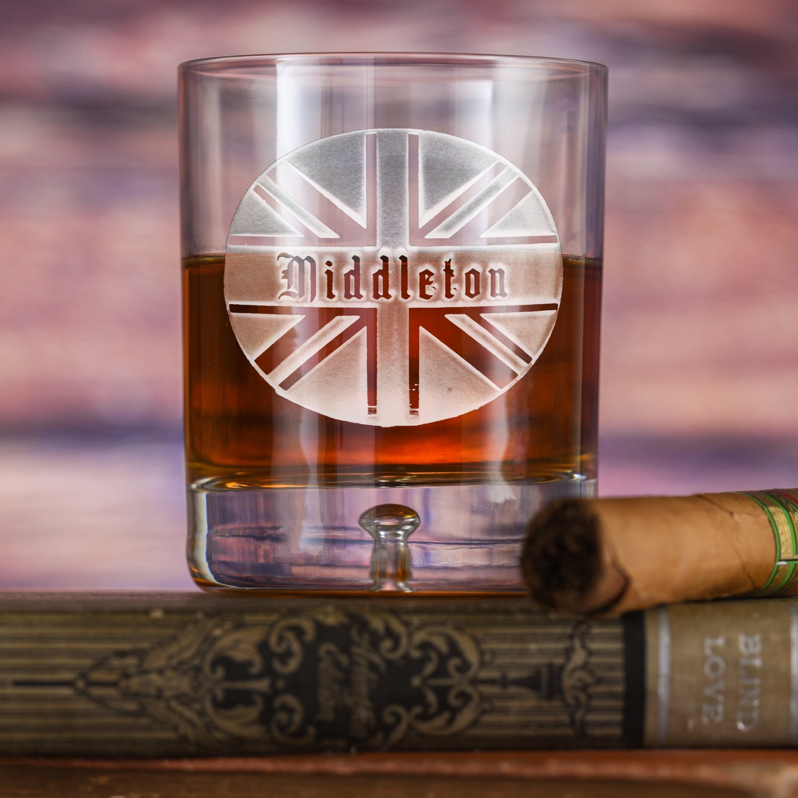 Clear rocks glass with a rounded base, filled with amber liquid, deep-carved with a Union Jack flag circle and the name "Middleton" in an old-English font, on a wood background.