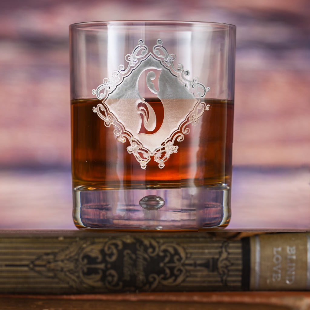 Rocks glass filled with amber liquid, deep-carved with a diamond-shaped ornate frame around a script letter "S," resting on a book.