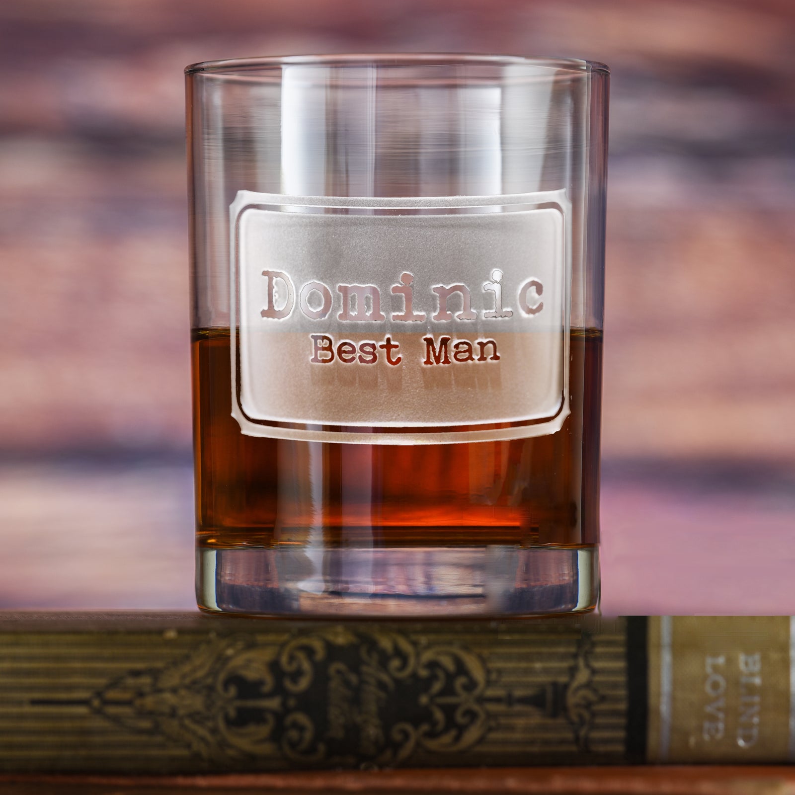 Rocks glass filled with amber liquid, deep-carved with a badge reading "Dominic, Best Man," resting on a book.