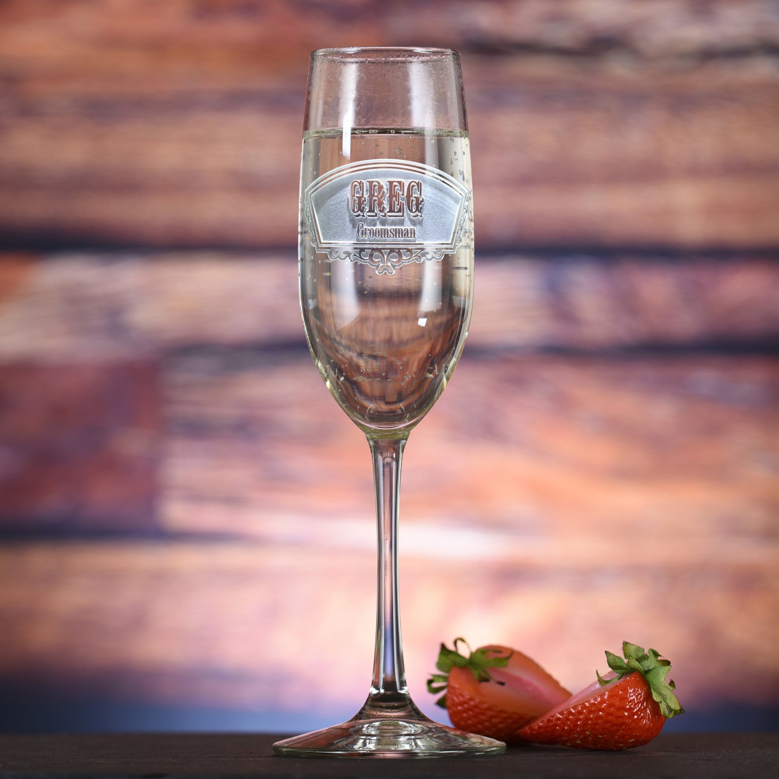 Champagne flute filled with champagne, engraved with a banner badge reading "Greg, Groomsman," on a sunset-toned wood background with strawberries.