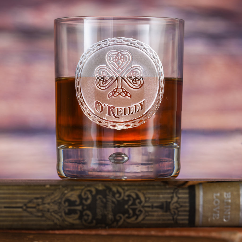 Clear rocks glass filled with amber liquid, deep-carved with a Celtic-knot shamrock above the name "O'Reilly," on a book.