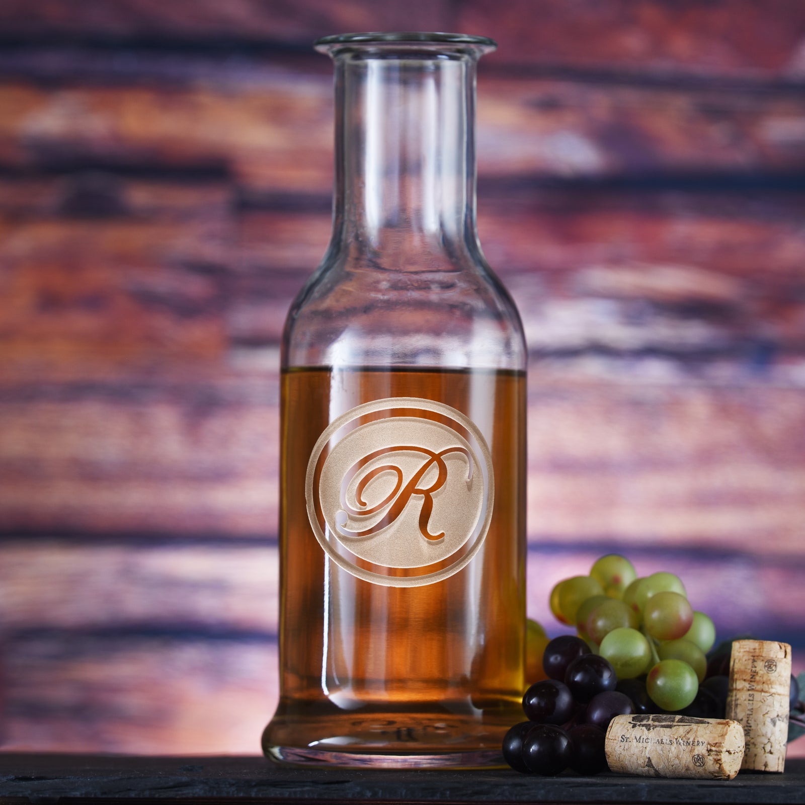Round-shoulder wine carafe filled with white wine, deep-carved with a single script initial inside a thin circle, styled with grapes.
