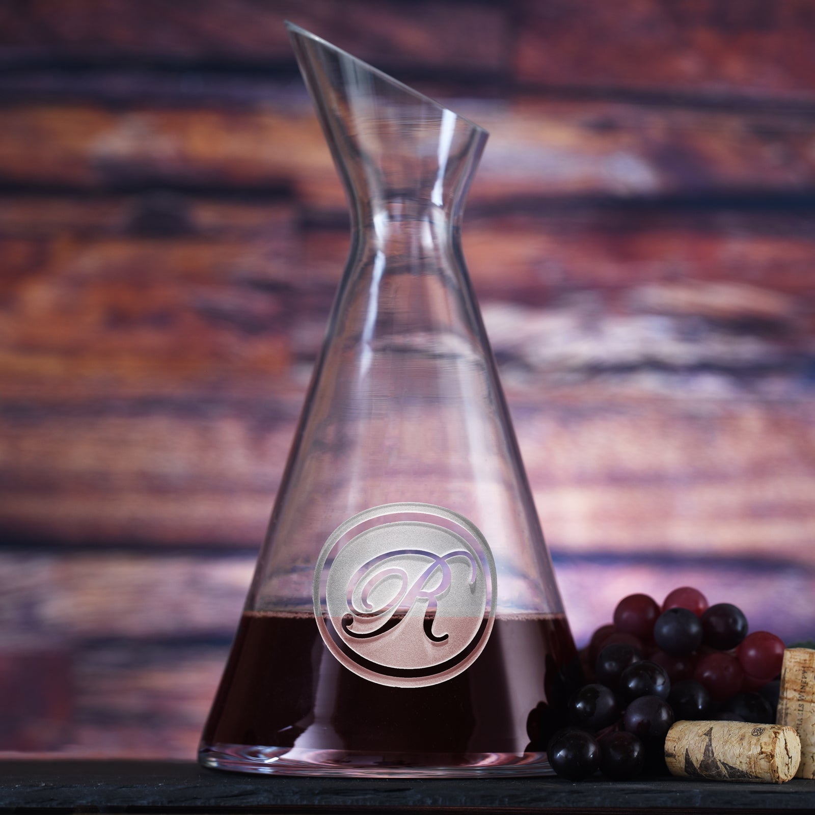 Slant wine carafe filled with red wine, deep-carved with a single script initial inside a thin circle, styled with grapes.