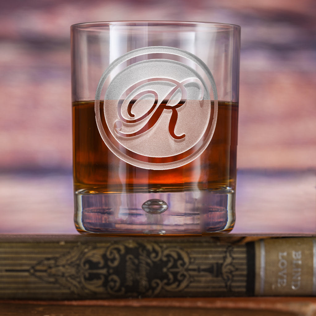 Rocks glass filled with amber liquid, deep-carved with a circular border around a script letter "R," on a wood background.