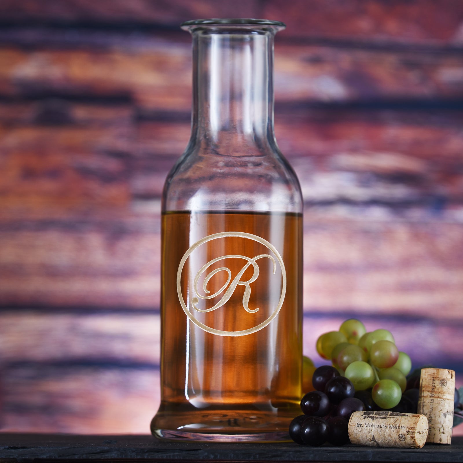 Round-shoulder wine carafe filled with white wine, deep-carved with a single script initial inside a thin circle, styled with grapes.