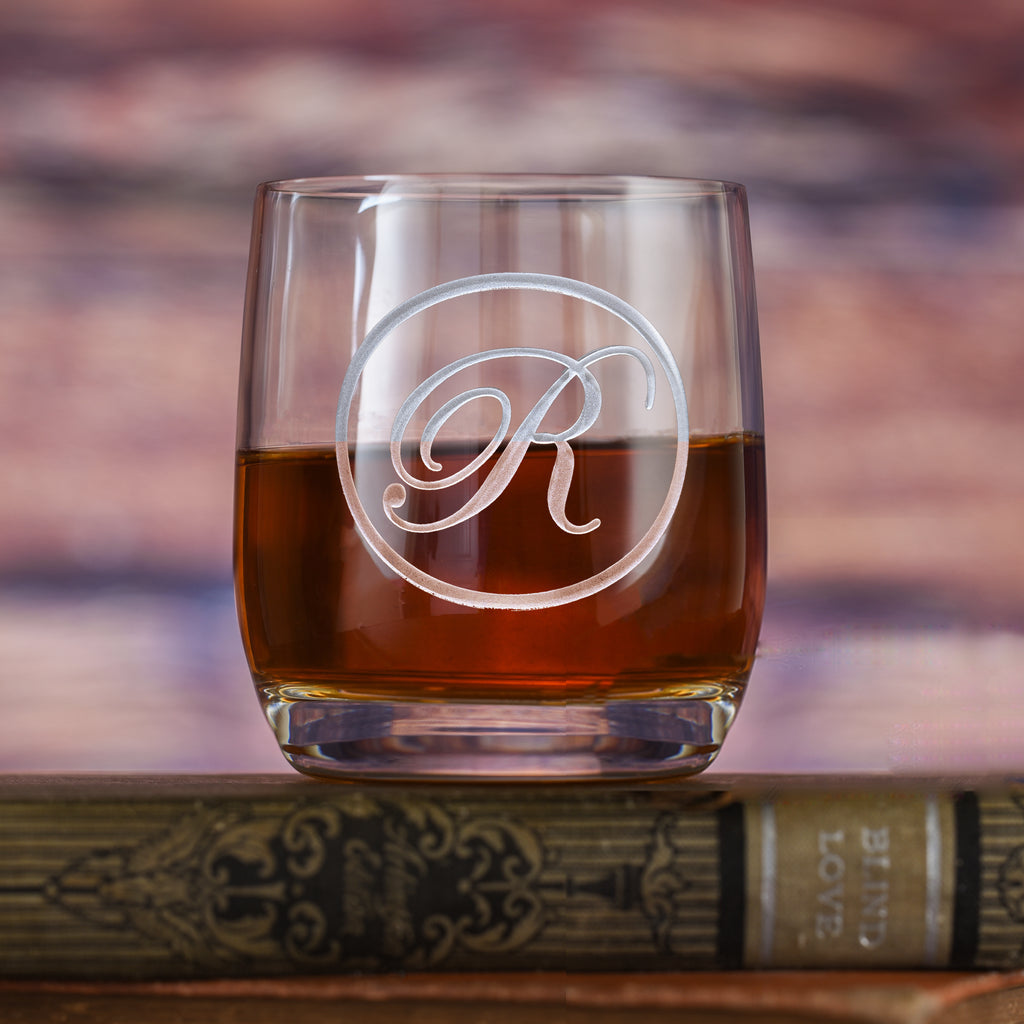 Rocks glass filled with amber liquid, deep-carved with a circular border around a script letter "R," resting on a book.