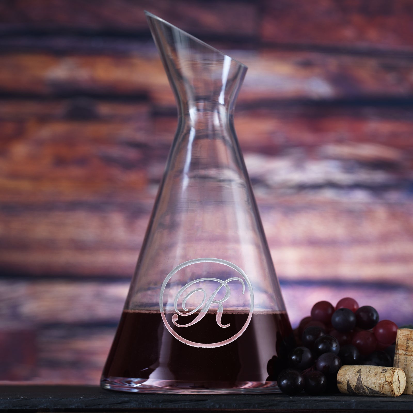 Slant wine carafe filled with red wine, deep-carved with a single script initial inside a thin circle, styled with grapes.