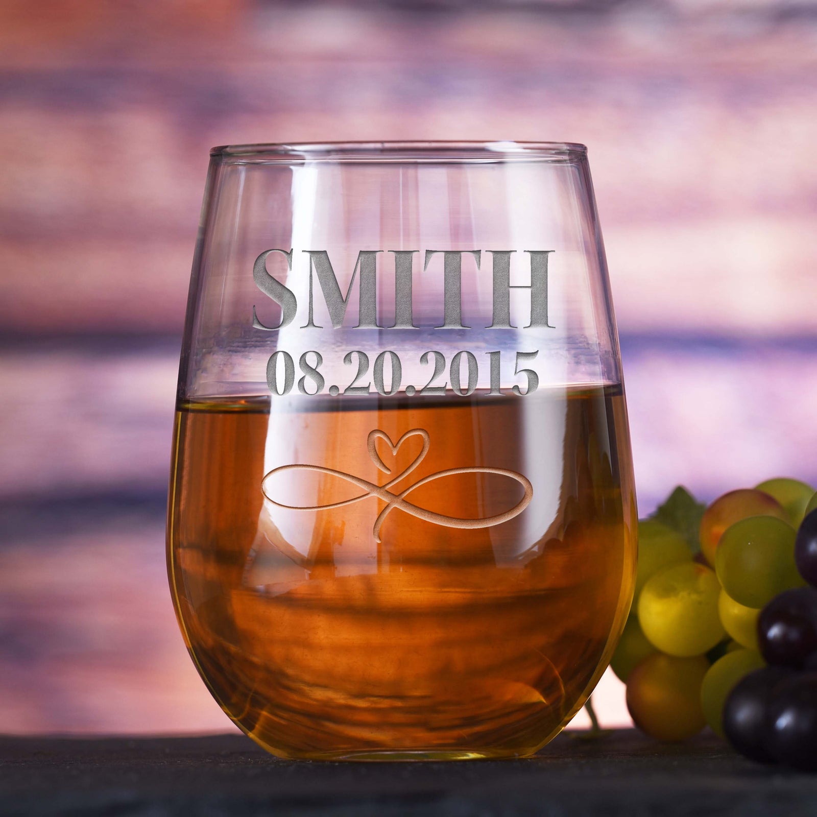 Stemless wine glass filled with white wine, deep-etched with the name "Smith," the date "08.20.2015," and an infinity-heart design, on a sunset-toned background with grapes.