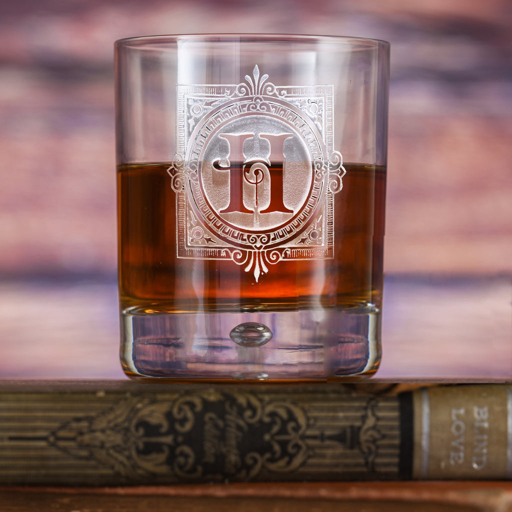 Rocks glass filled with amber liquid, deep-carved with a shield reading "H, Harrison," on a wood background.
