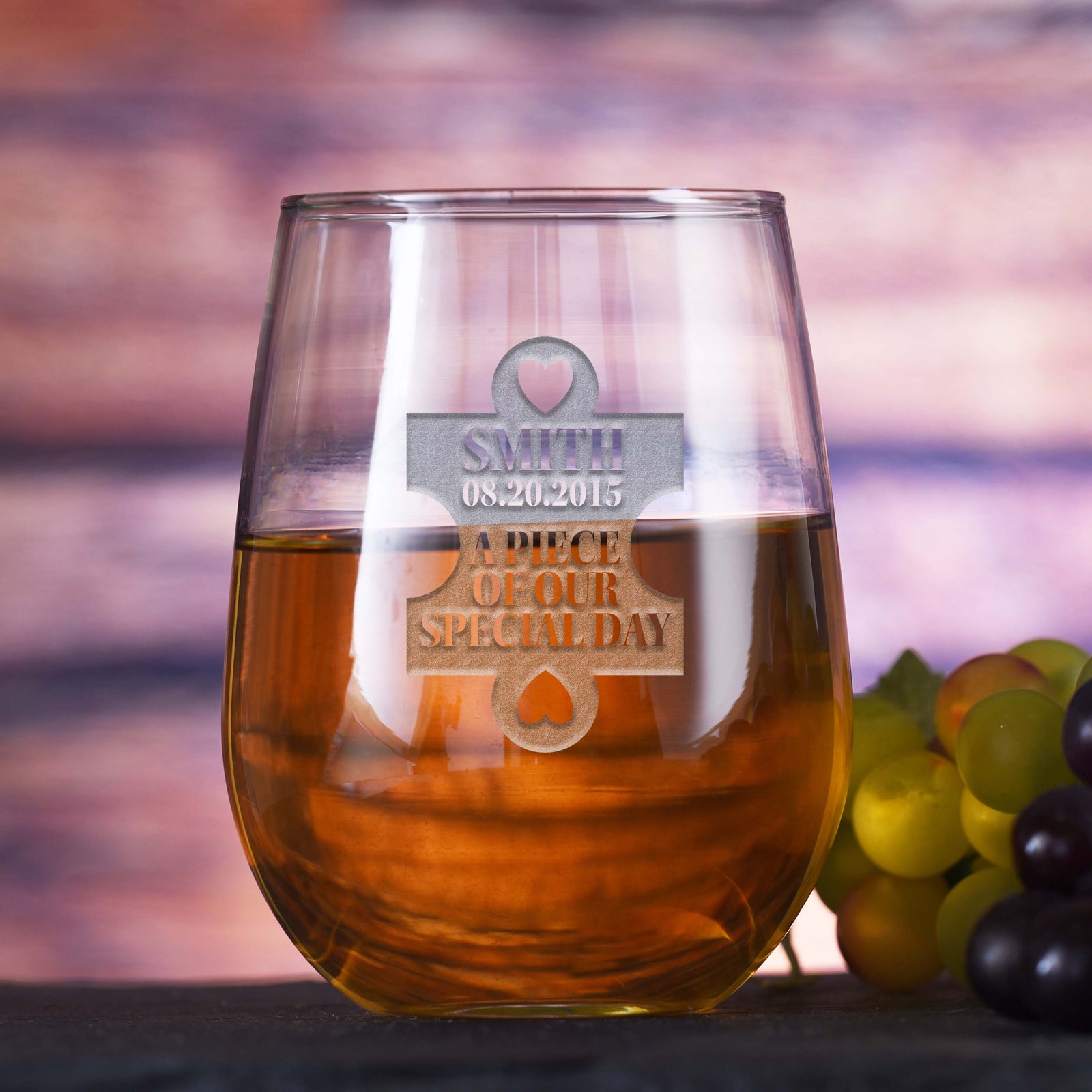 Stemless wine glass filled with white wine, deep-etched with a puzzle-piece design reading "Smith, 08.20.2015, A Piece of Our Special Day," on a sunset-toned background with grapes.