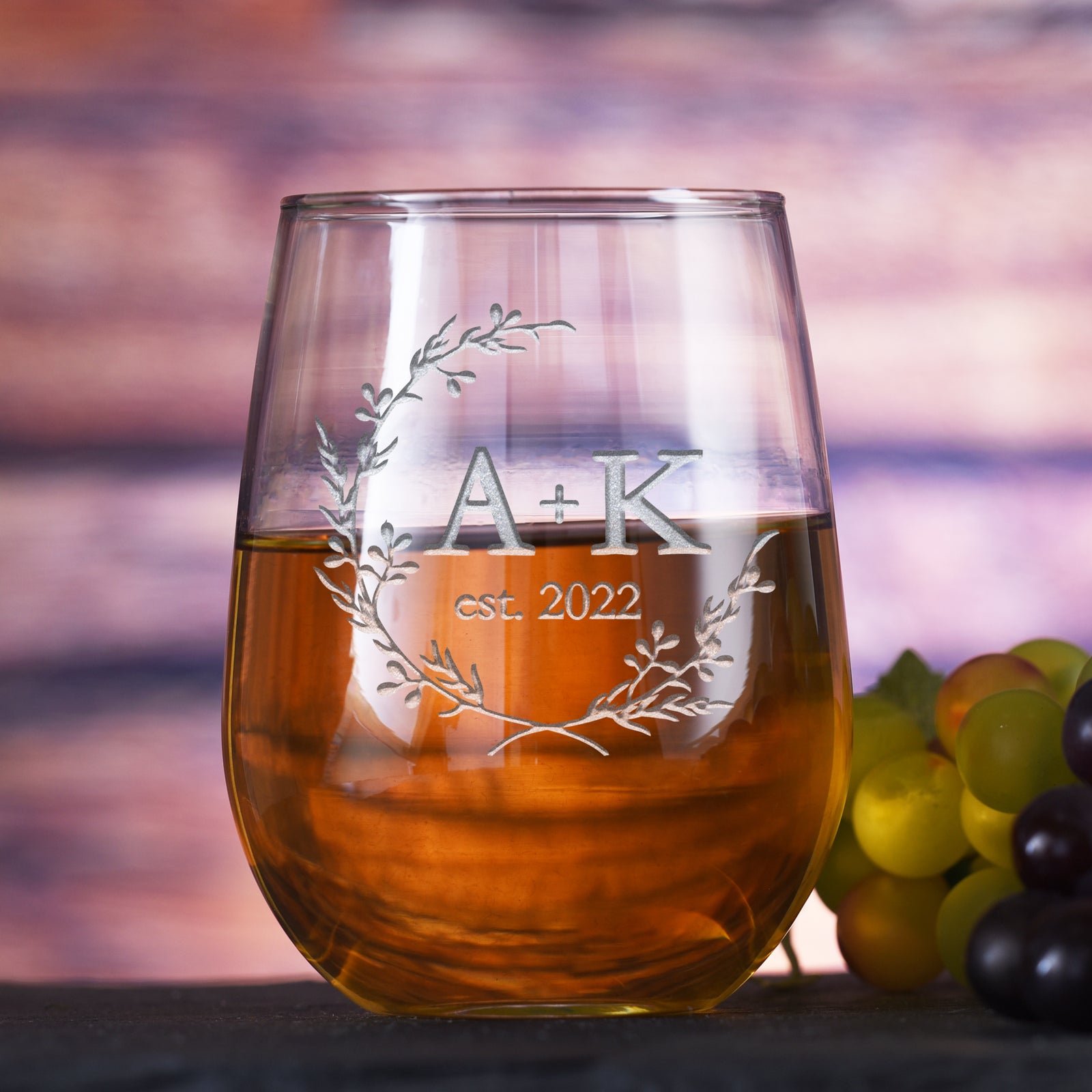 Stemless wine glass filled with white wine, deep-etched with a laurel wreath around the initials "A + K" and the text "est. 2022," on a sunset-toned background with grapes.