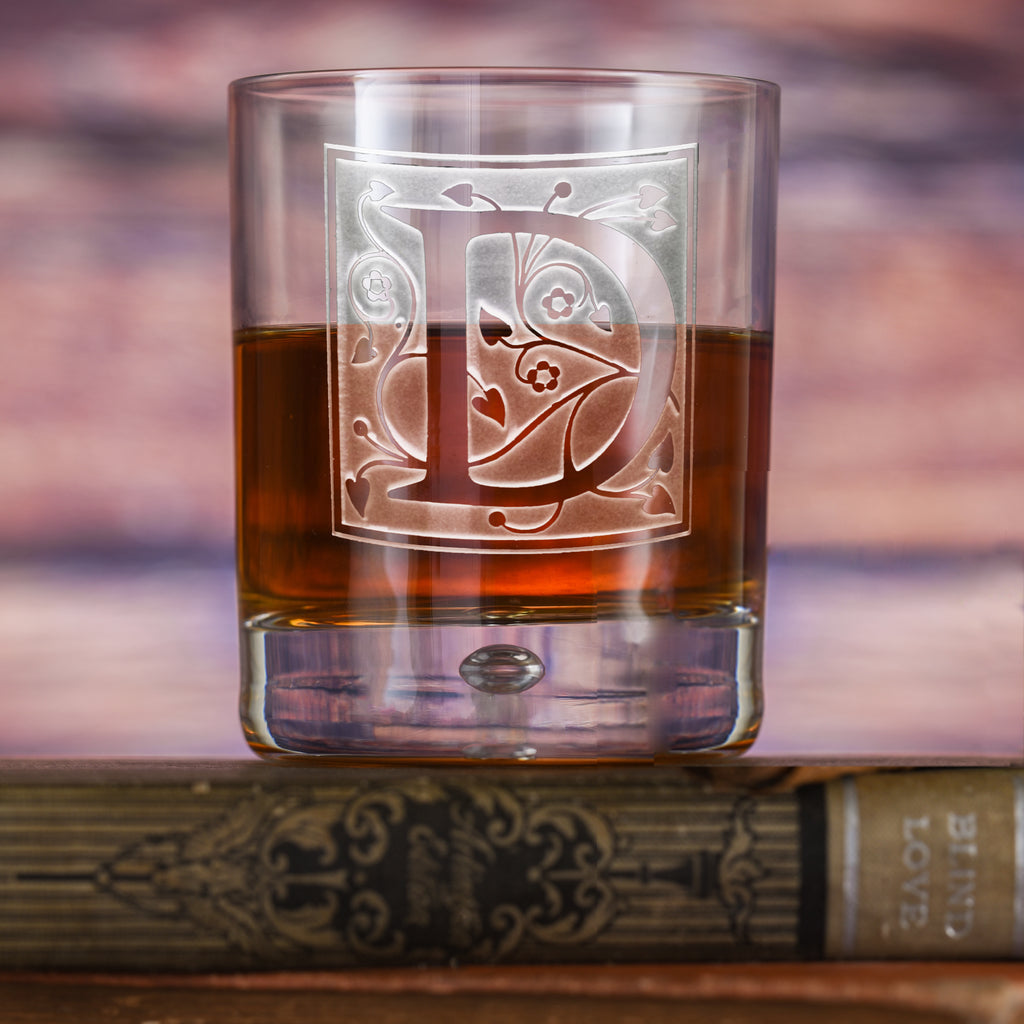 Rocks glass filled with amber liquid, deep-carved with an ornate letter "D" framed by a floral and heart vine pattern, resting on a book.
