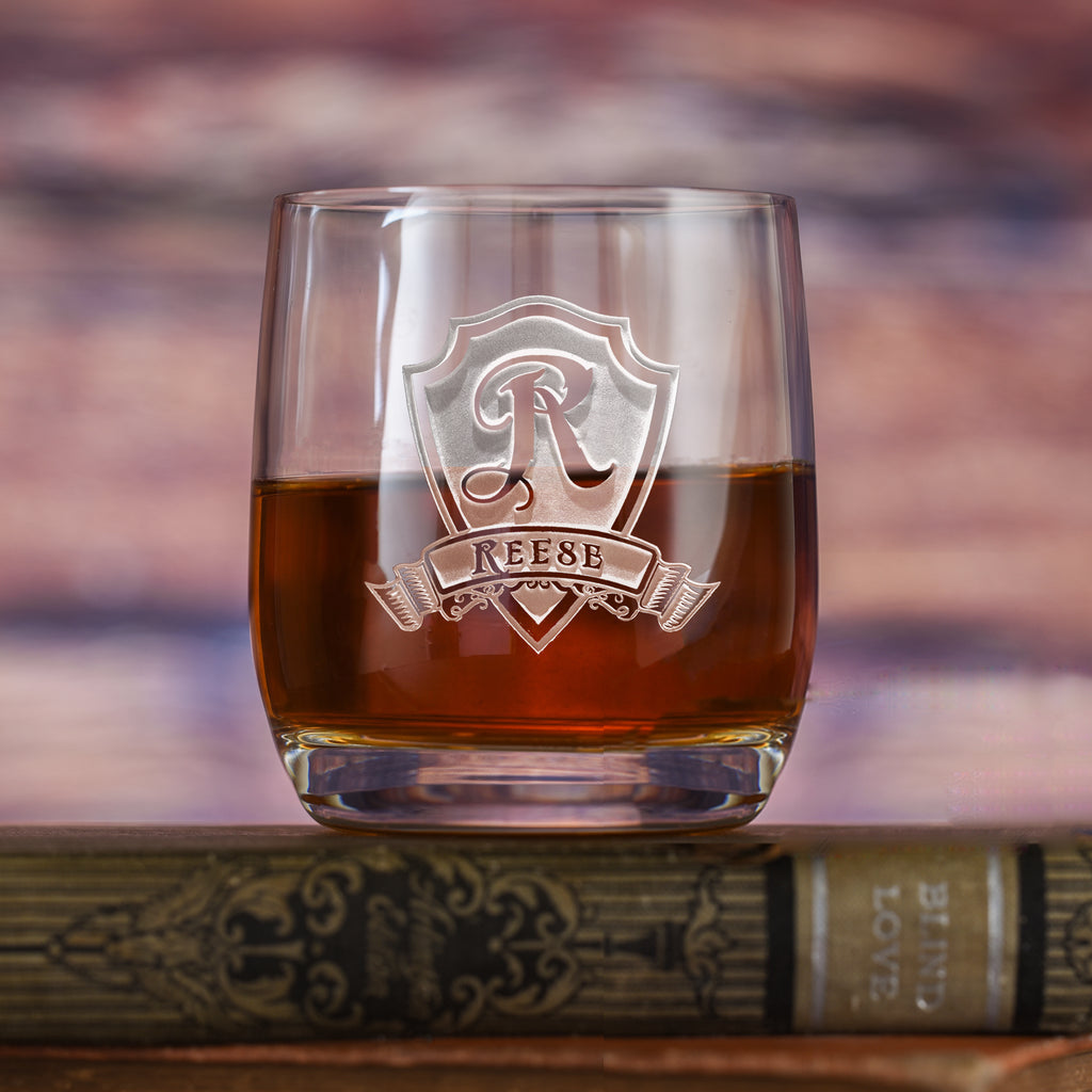 Rocks glass filled with amber liquid, deep-carved with a shield around the letter "R" with a banner reading "Reese," resting on a book.