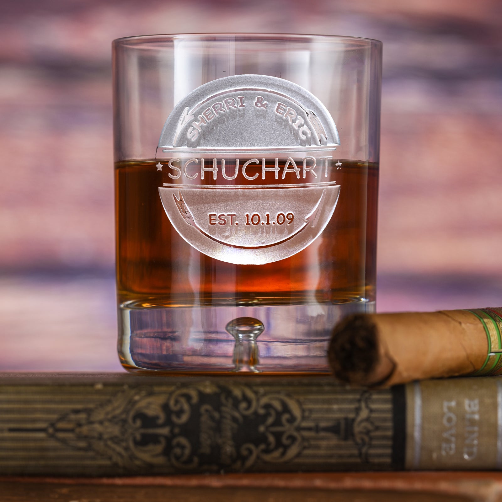 Rocks glass filled with amber liquid, deep-carved with a wedding badge reading "Sherri & Eric, Schuchart, Est. 10.1.09" with arrow motifs, resting on a book with a cigar.