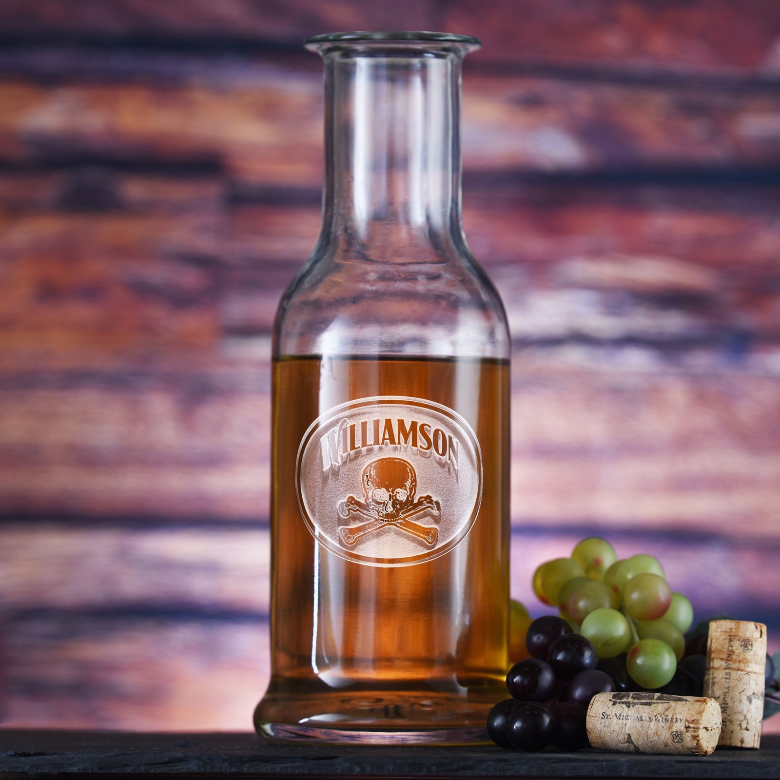 Round-shoulder wine carafe filled with white wine, deep-carved with the name "Williamson" above a skull and crossbones in an oval, styled with grapes.