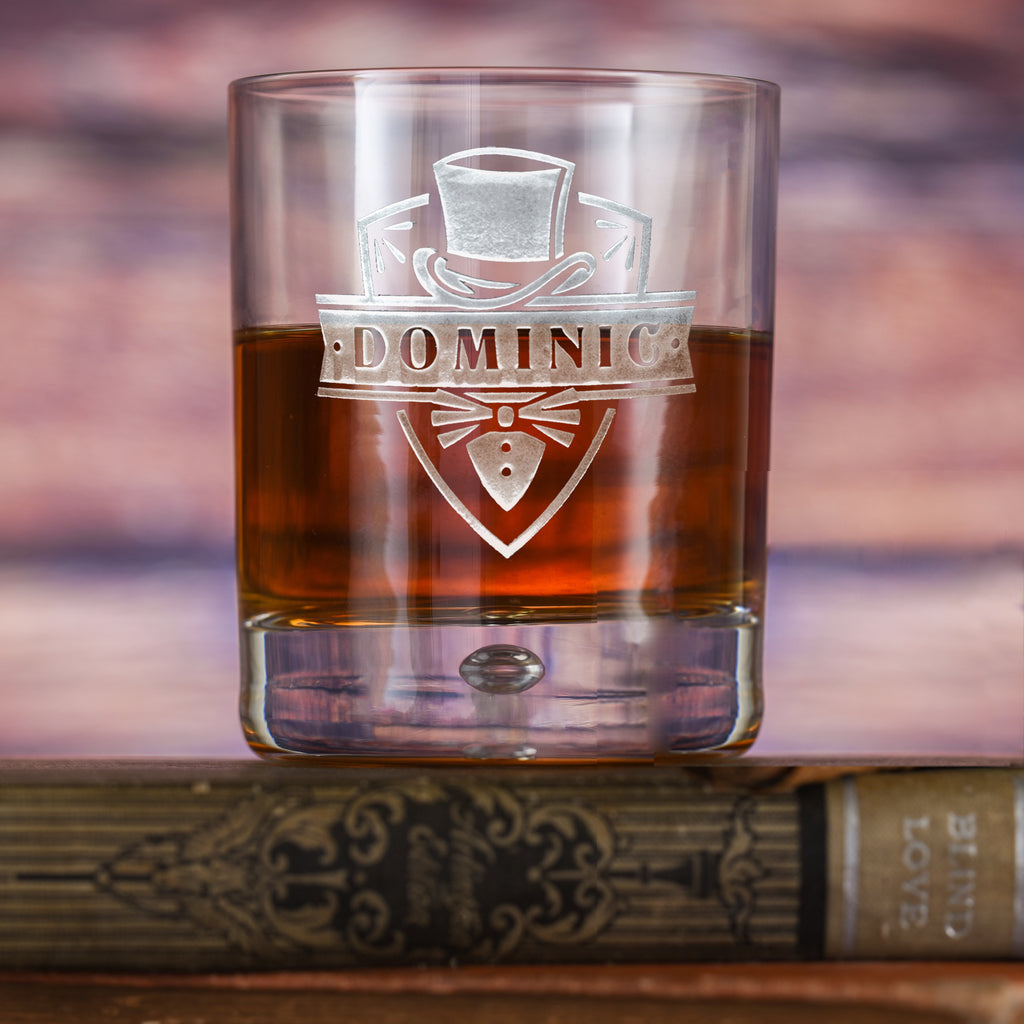 Rounded-base rocks glass filled with amber liquid, deep-carved with a shield reading "Dominic" below a top hat and bowtie, resting on a book.