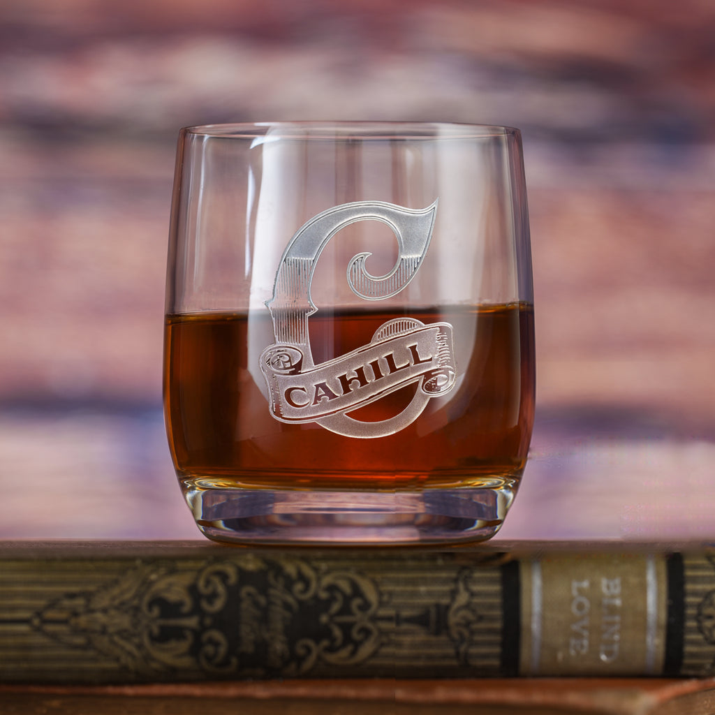 Clear rocks glass filled with amber liquid, deep-carved with a shield reading "C, Cahill," on a book background.
