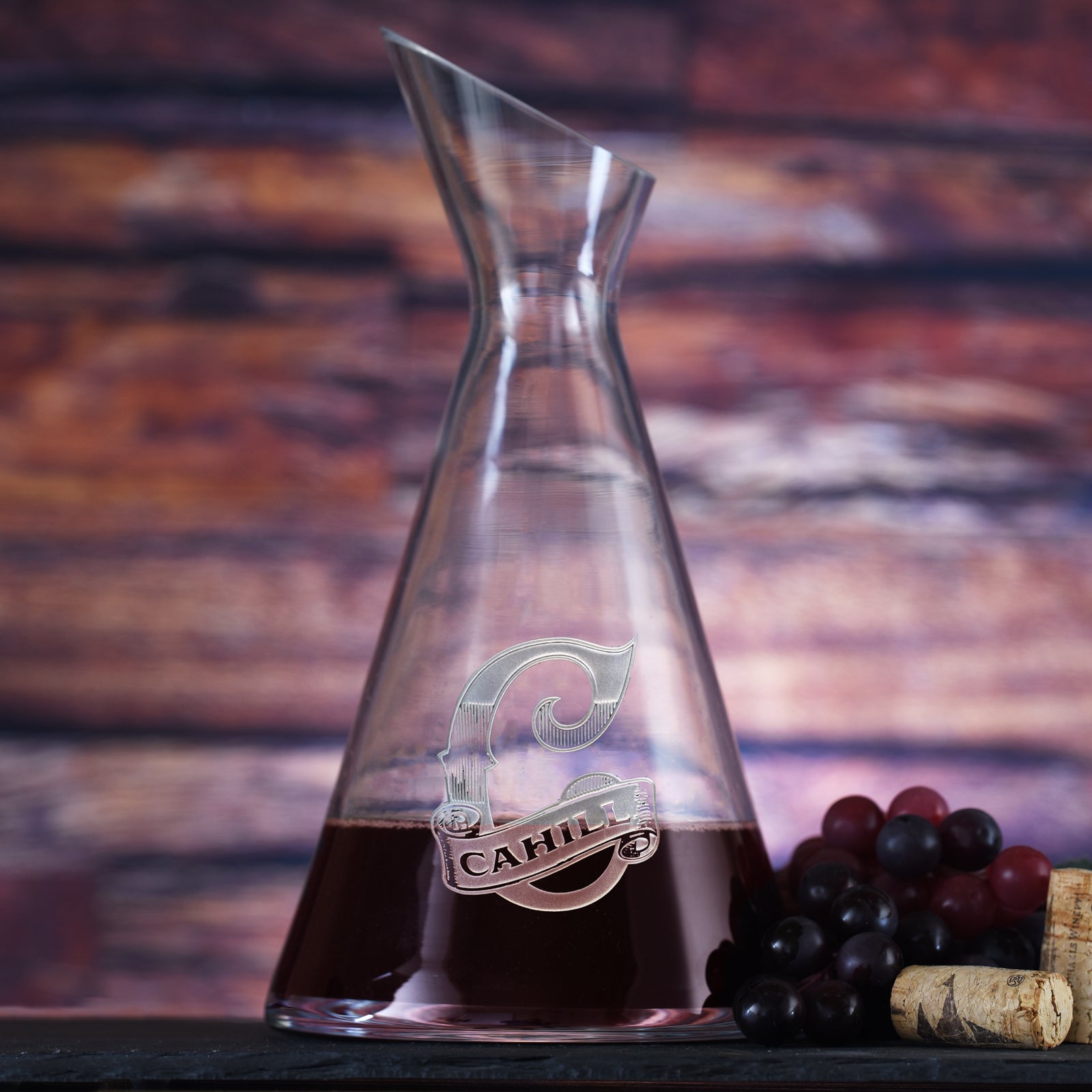Slant wine carafe filled with red wine, deep-carved with a script initial and a last name, styled with grapes.