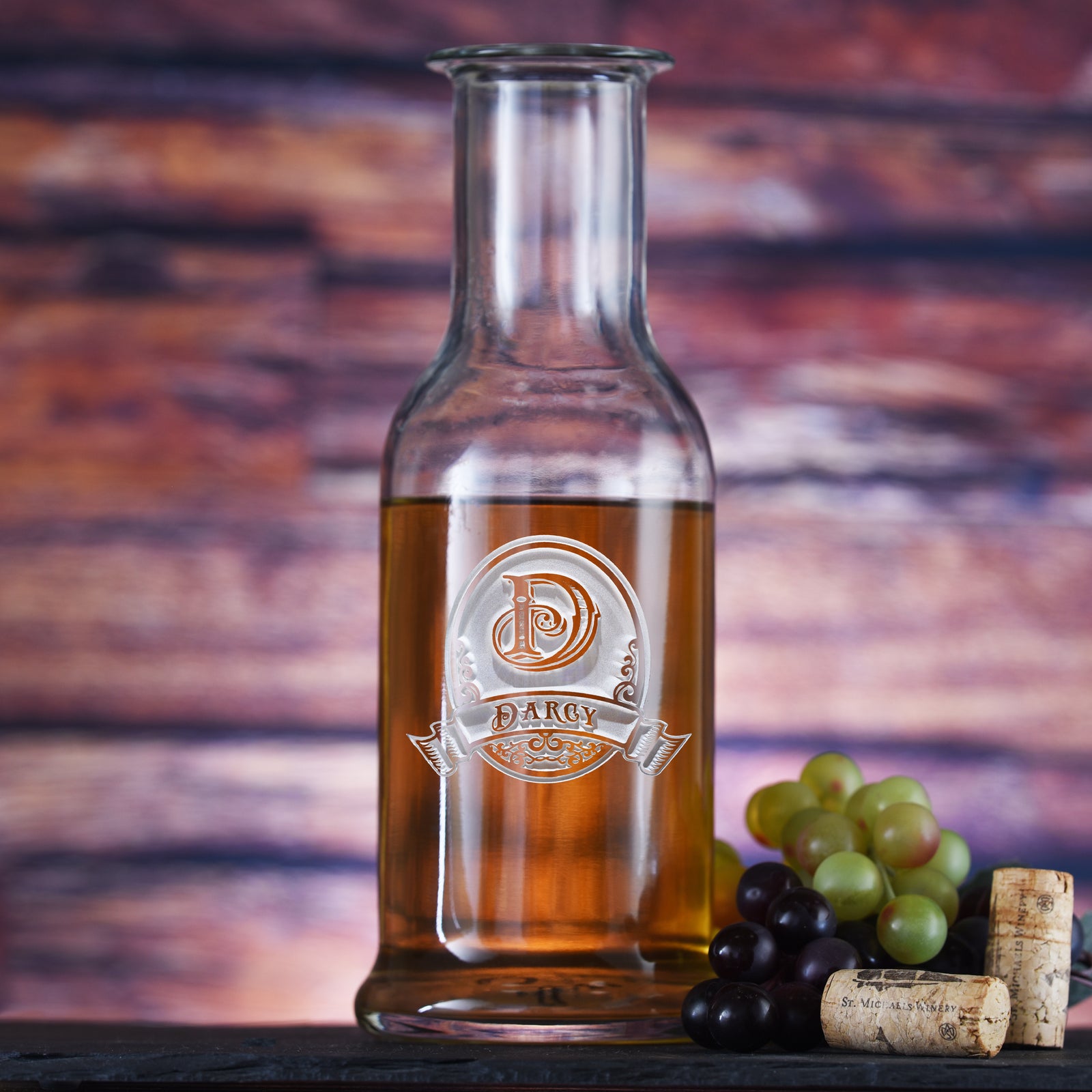 Glass wine carafe filled with amber liquid, deep-carved with an oval crest reading "D, Darcy," on a wood background with grapes and corks.