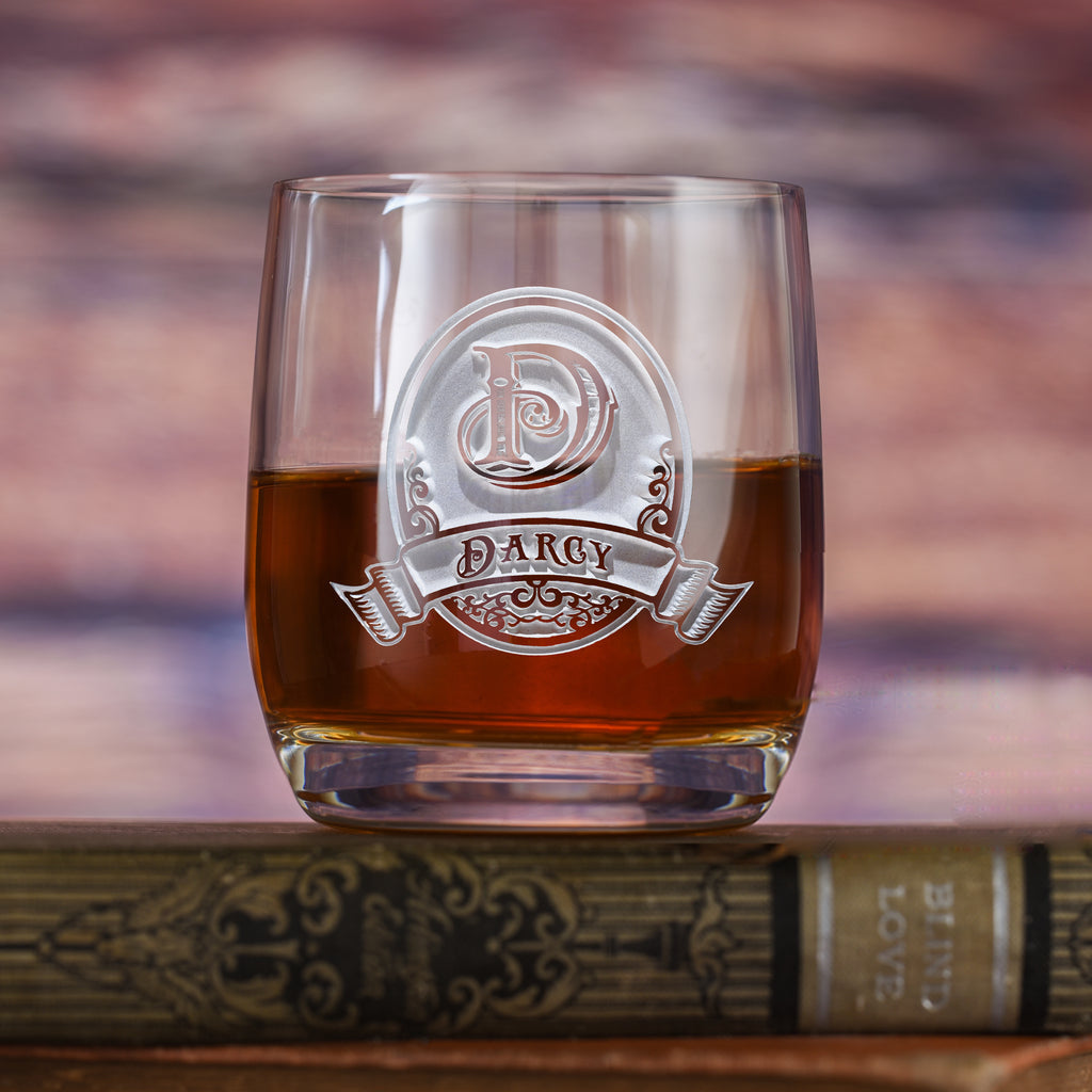 Rocks glass filled with amber liquid, deep-carved with an oval crest reading "D, Darcy," resting on a book.