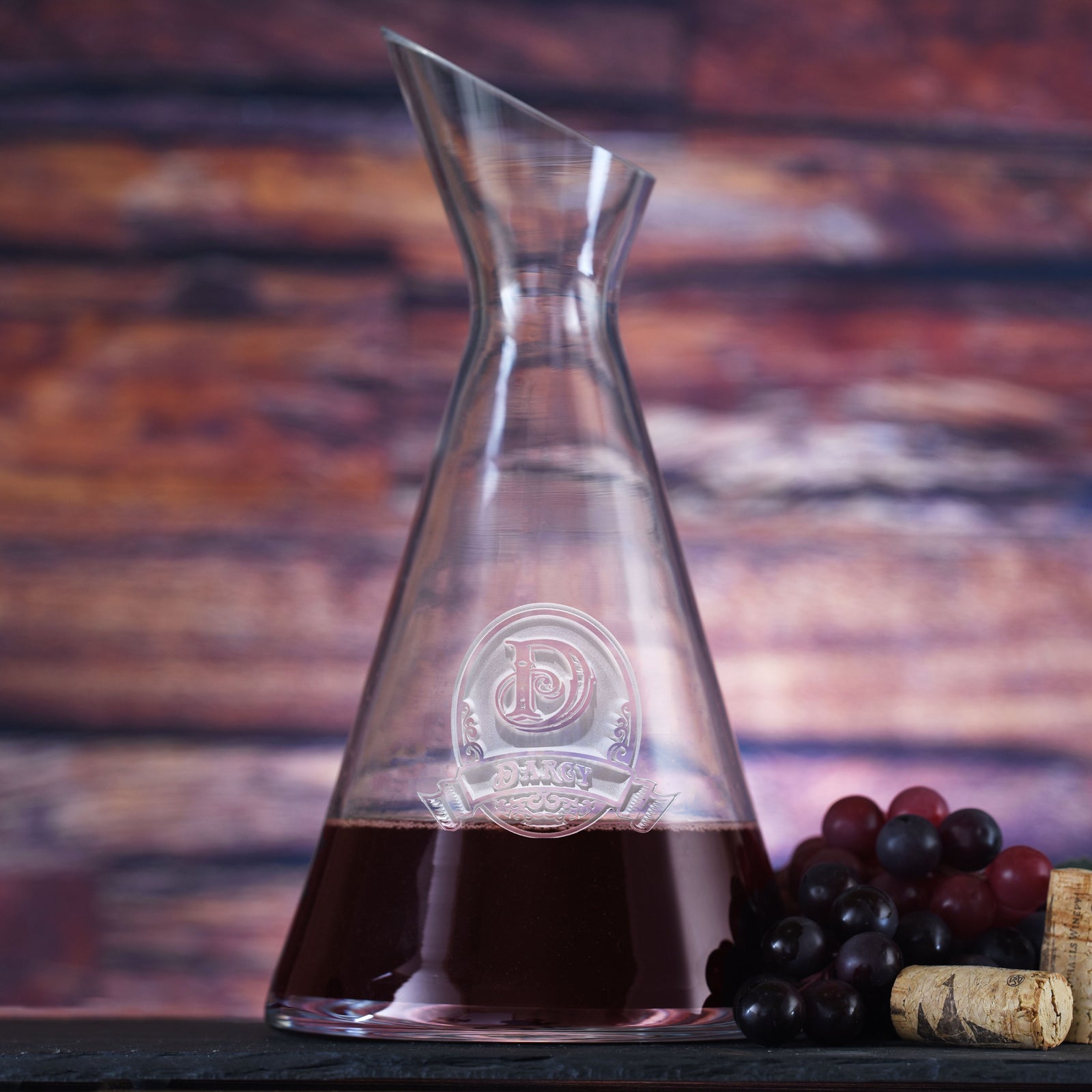 Slant wine carafe filled with red wine, deep-carved with an initial in an oval crest above a name banner, styled with grapes.