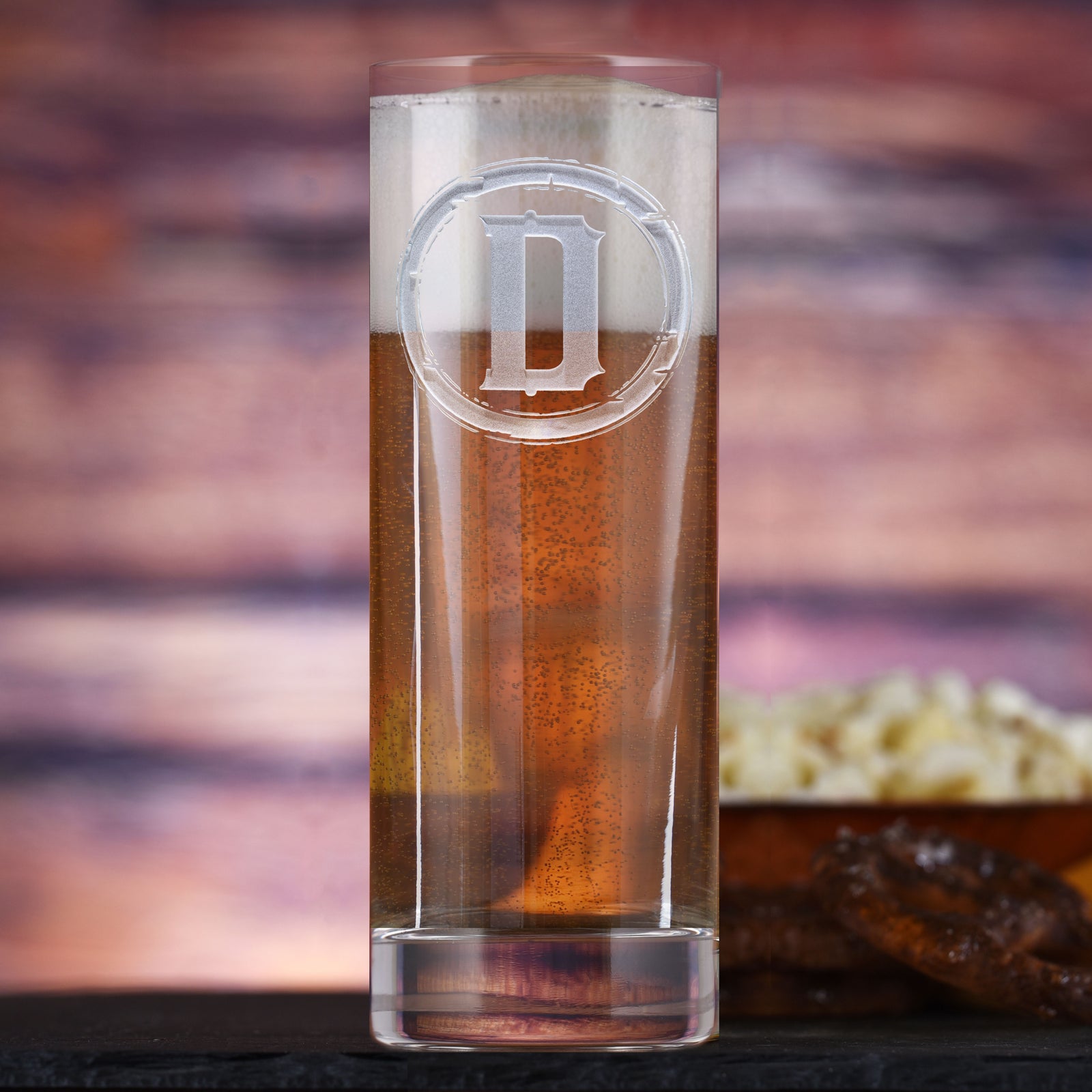 Tall New York tumbler filled with beer, deep-carved with a distressed circular border around the letter "D," on a wood background with a bowl of snacks.