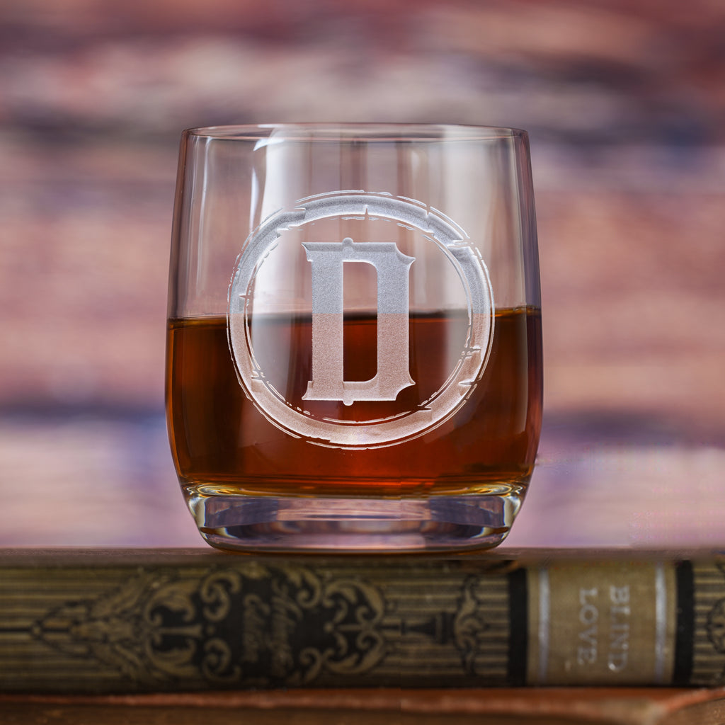 Rocks glass filled with amber liquid, deep-carved with a distressed circular border around the letter "D," resting on a book.