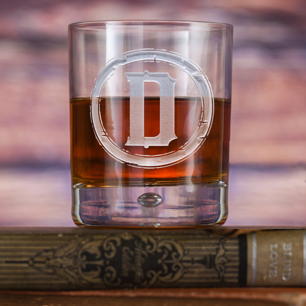 Rocks glass filled with amber liquid, deep-carved with a distressed circular border around the letter "D," resting on a book.