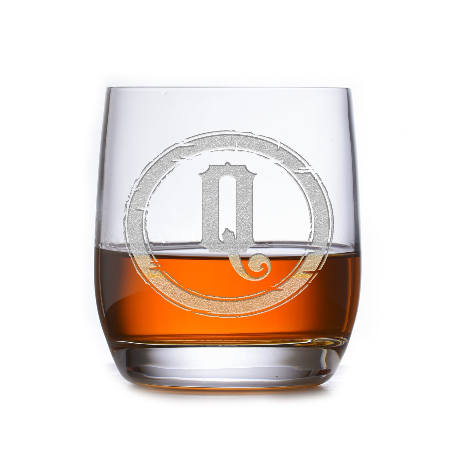 Rocks glass filled with amber liquid, deep-carved with a distressed circular border around the letter "Q," on a white background.