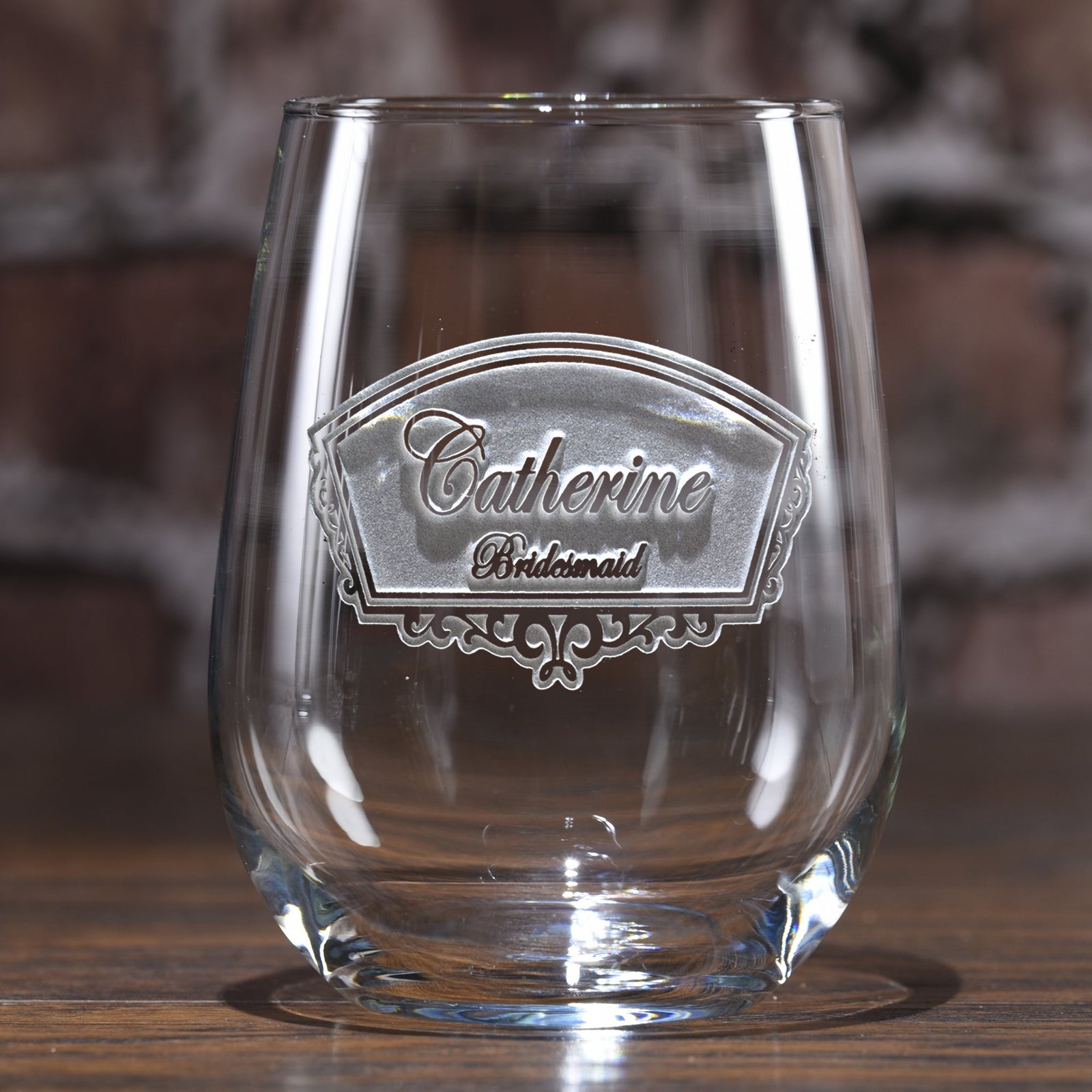 Stemless wine glass deep-etched with an ornate frame reading "Catherine, Bridesmaid," on a blurred wood-toned background.