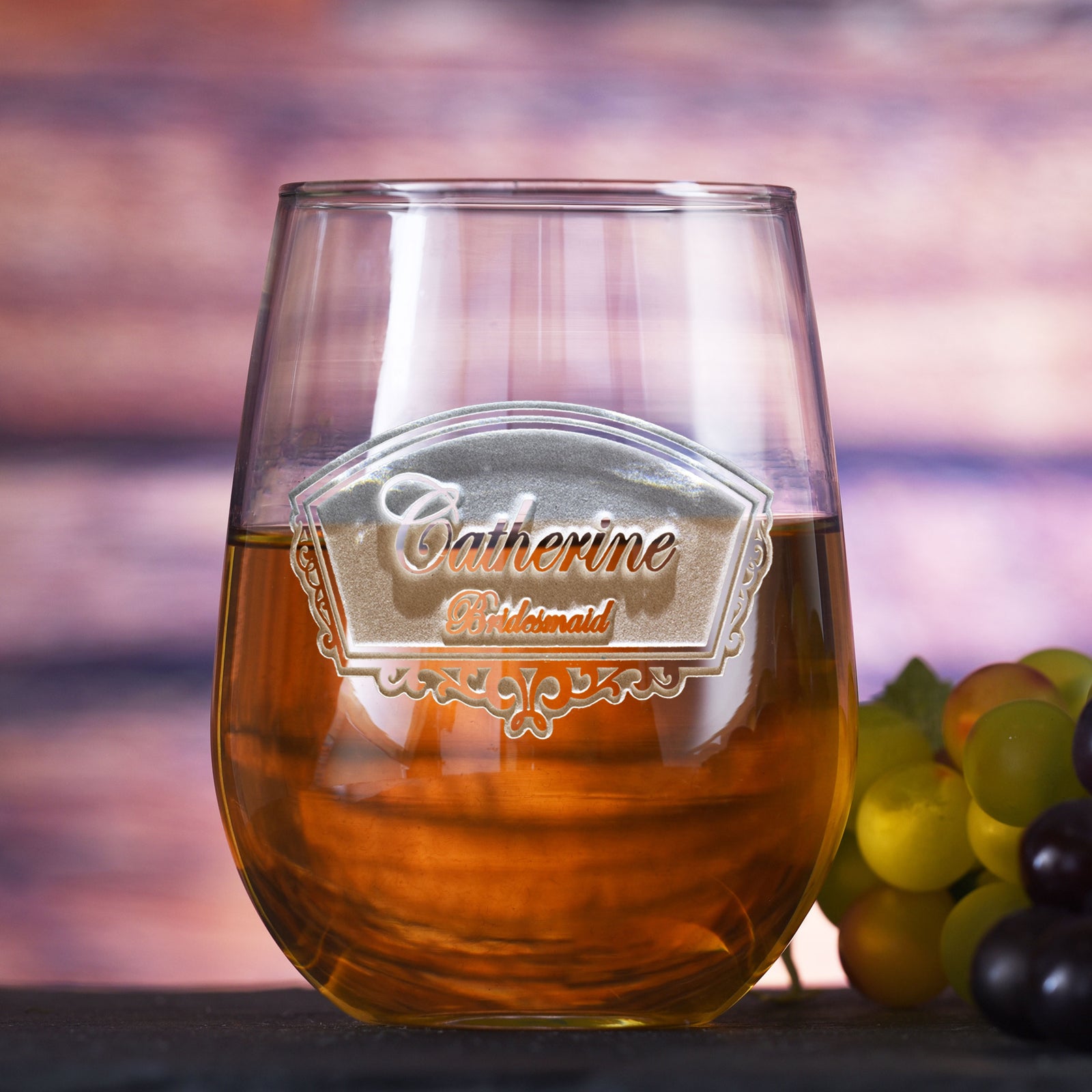 Stemless wine glass filled with amber wine, deep-etched with an ornate frame reading "Catherine, Bridesmaid," on a sunset-toned background with grapes.