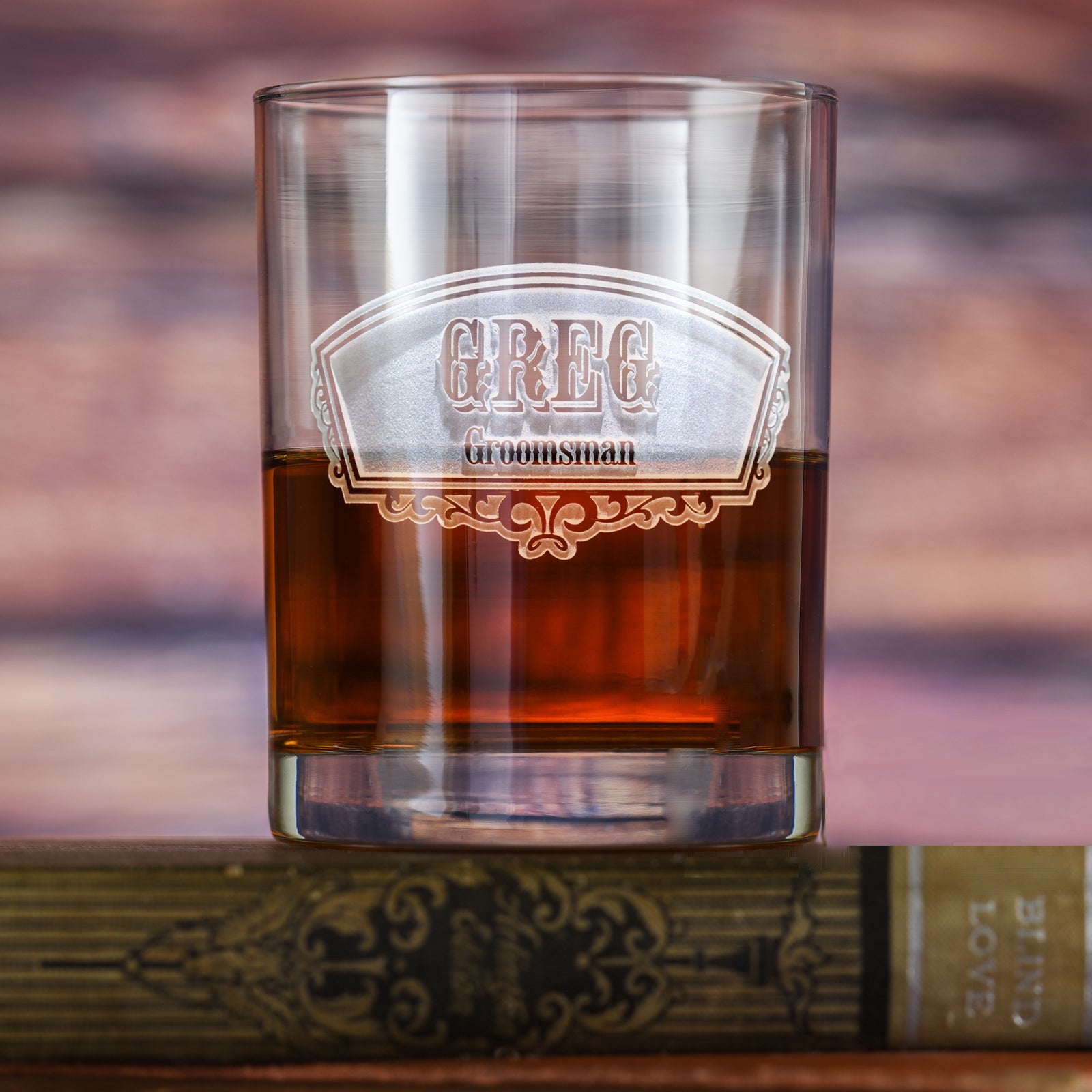 Tapered rocks glass filled with amber liquid, deep-carved with a fan-shaped badge reading "Greg, Groomsman," resting on a book.