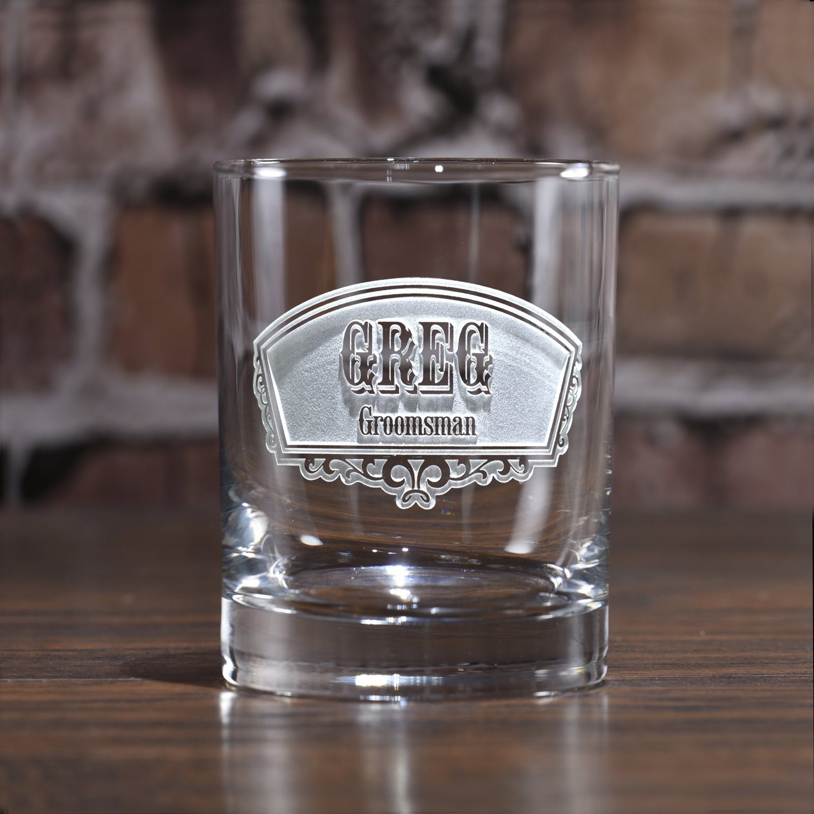 Tapered rocks glass deep-carved with a fan-shaped badge reading "Greg, Groomsman," on a brick background.