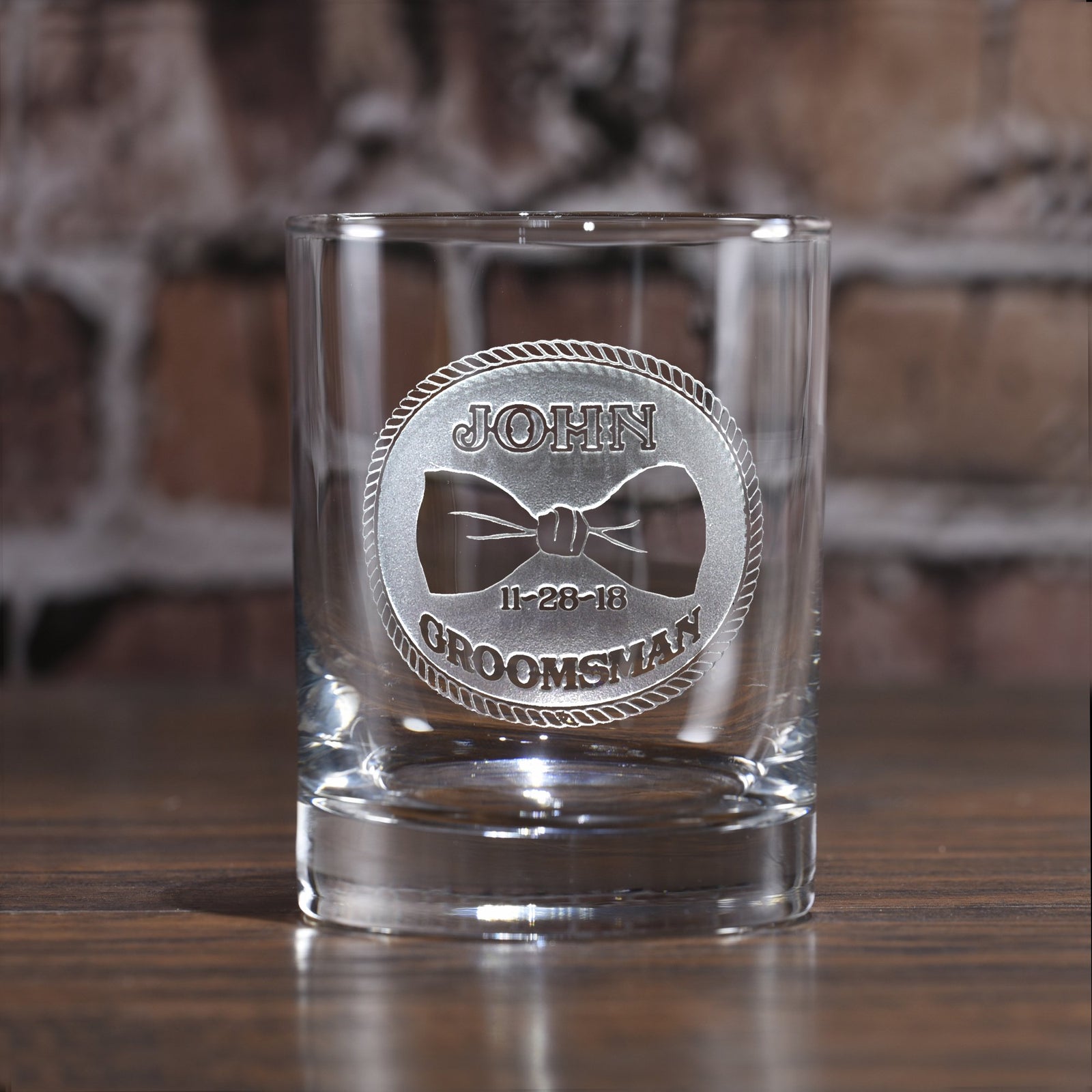 Rocks glass deep-carved with a circular badge reading "John, Groomsman, 11-28-18" around a bowtie, on a brick background.