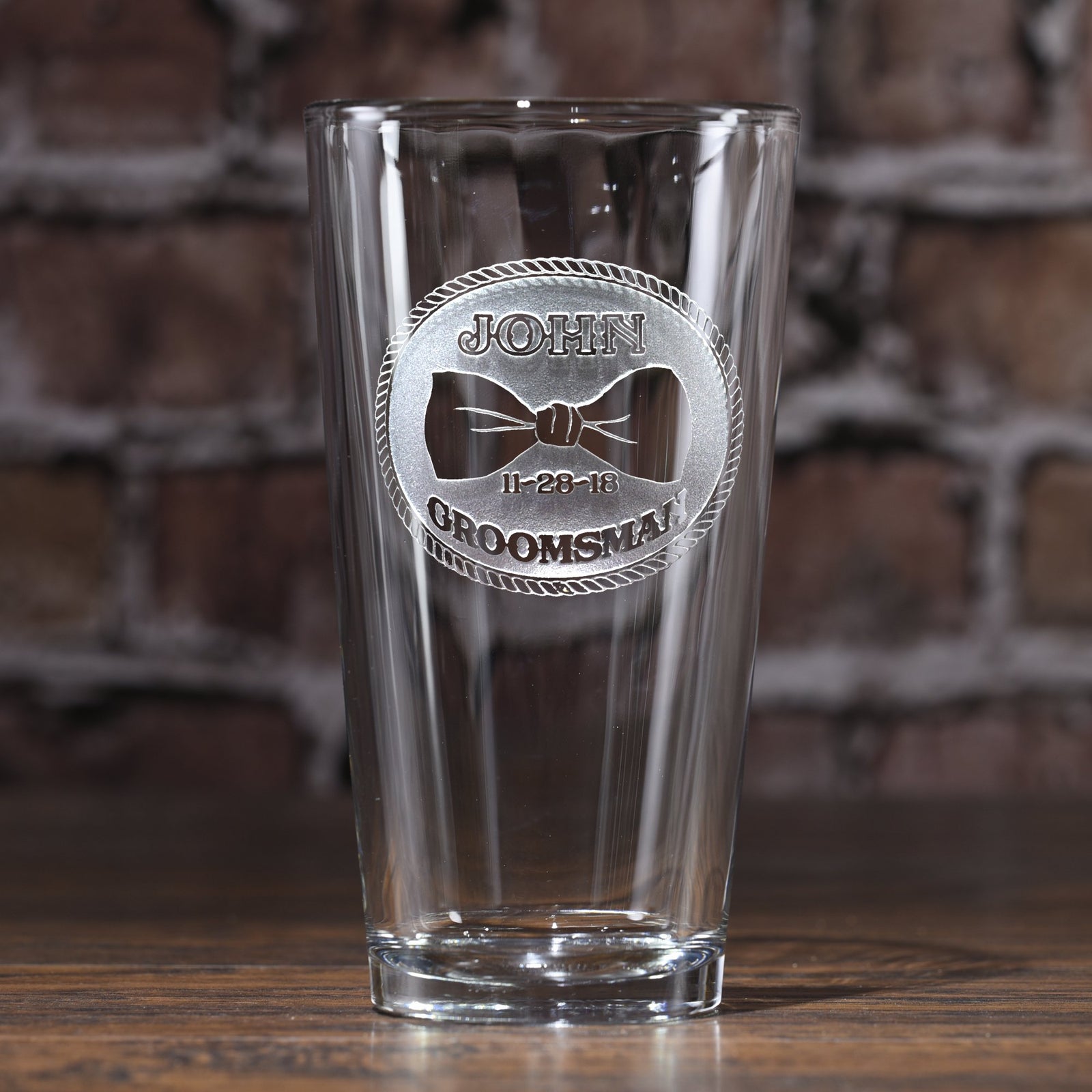 Empty pint beer glass deep-etched with a rope-bordered circular badge around a bowtie, reading "John, 11-28-18, Groomsman," on a blurred brick background.