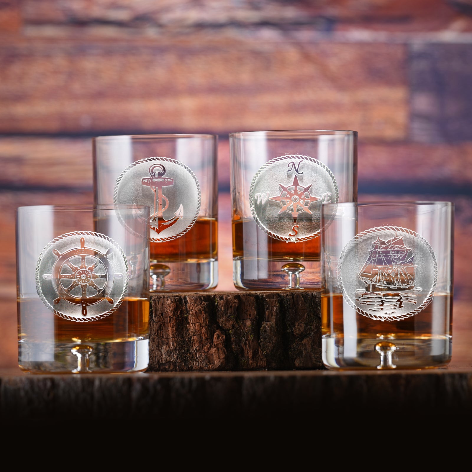 Four rocks glasses filled with amber liquid, each deep-carved with a roped nautical medallion (anchor, ship's wheel, compass rose or tall ship), on a wood-log background.